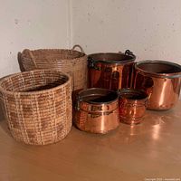 View of 4 copper buckets and 2 woven baskets on a wooden surface against a textured wall