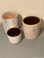Three ceramic crocks displayed, including a large beige crock with a dark brown interior and crown 3 stamp, a smaller ceramic crock, and a textured beige pot.