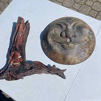 Photo of a metal wall hanging shaped as a smiling moon face along with a wooden carving featuring a smiling face integrated into a piece of wood, both placed on a white surface.