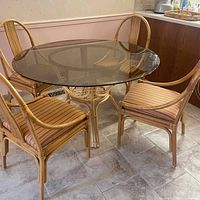 Photo showing round glass-top dining table with four rattan chairs, visible tile floor and kitchen corner.