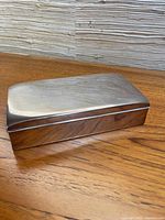 Closed rectangular silver cigarette box placed on wooden surface showing smooth polished engraved lid and sides.