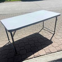 Full view of the folding utility table fully set up outdoors on a paved surface, showing white top and metal legs.