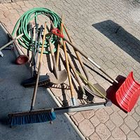 Photo showing entire lot of garden hose coiled with black connectors alongside various yard tools including a broom, rake, shovel, hoe, and plastic snow shovel.