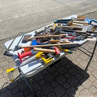 Assorted hardware tools spread on a white foldable table outside on pavement under sunlight.