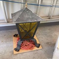 Metal framed exterior lantern light with yellow tinted etched glass panels on a wooden board with orange extension cord underneath.