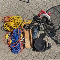 Full view of all items in lot on pavement: circular saw, angle grinder, impact drill, three extension cords, and knee pads.