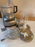 Full view of KitchenAid food processor base, clear plastic processing bowl and lid with chute, two stainless steel discs, plastic container, power cord visible