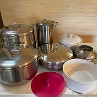 Full view of stainless steel pots, ceramic bowls with lids, red plastic lid, and steamer basket.