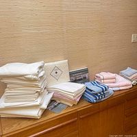 Stack of assorted folded linens including white sheets, blue striped towels, pink towels, and neutral cloths on wooden cabinet.