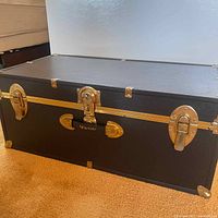Front view of a black Seward trunk with gold-tone lock and handle, showing the overall shape and condition.