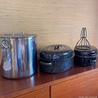 Wide shot showing stainless steel stock pot, two black speckled enamel roasting pans with lids, and a metal whisk resting on the smallest pan.