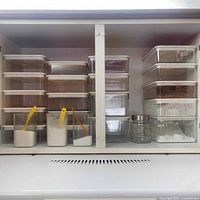 Wide cabinet shelf showing stacked clear rectangular plastic storage containers with white lids holding various dry ingredients. Three clear square containers with plastic scoops and small glass jar also visible.