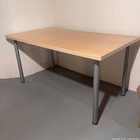 Full view of rectangular utility table with light wood top and silver metal legs placed against a wall on a gray floor.