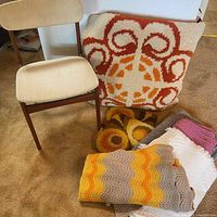Photo shows vintage teak chair with beige cushioned seat and backrest beside multiple retro hanging rugs and textiles with vibrant orange, cream, brown, yellow, and pink patterns.