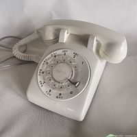 Off-white vintage rotary dial telephone with handset resting on cradle. Coiled cord visible. Dial includes alphanumeric codes.