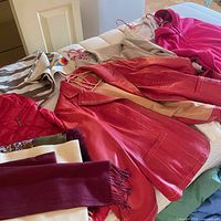 Assorted women's clothing laid on bed, including red leather jacket, red puffer coat, beige coat, pink garment, and scarves.