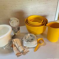 Wide angle photo showing full set of yellow measuring bowls nested, yellow pitcher, white kitchen scale, reusable piping bag, plastic squeeze bottle with red cap, white mixing bowl with strainer insert, and small orange measuring strainer, all arranged on a white surface.