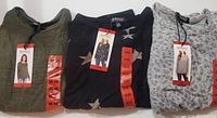 Three folded ladies knit long sleeve tops by Buffalo David Bitton, with visible labels and size medium tags, showing an olive green top, a black top with star prints, and a gray floral top.