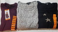 Three folded knit long sleeve tops laid side by side: dark burgundy, light gray heathered pattern, black with star applique.