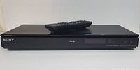 Front view of Sony Blu-Ray player with remote control placed on top, showing Sony logo and Blu-Ray symbol.