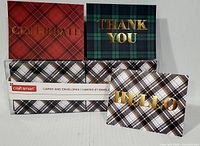 Three displayed note cards with plaid patterns and gold foil text 'Celebrate', 'Thank You', and 'Hello', along with the boxed set.
