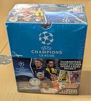 Front of the sealed box showing UEFA Champions League logo, Topps branding, and images of football players, along with guarantee of shiny stickers inside.
