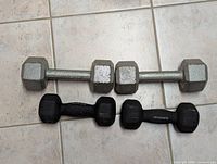 Photo of four dumbbell weights including two silver hexagonal metal dumbbells and two smaller black coated dumbbells on tiled floor.