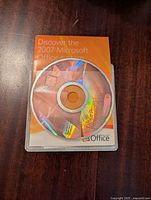 Microsoft Office Project Professional 2007 installation CD in clear plastic sleeve on wooden surface with orange manual booklet underneath.