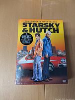 Front view of unopened Starsky & Hutch The Complete First Season 5-disc DVD set sealed in original shrink wrap. Shows the two main characters standing in front of a red and white coupe car with city skyline background.