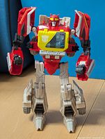Front view of Transformers Blaster figure showing red and gray color, yellow chest window with Autobot symbol, and arm and leg details.
