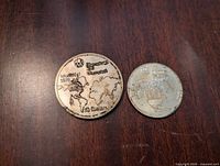 Both coins side by side on wooden surface showing the $10 and $5 coins with map design and inscriptions.