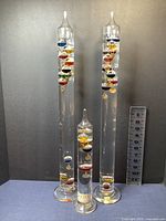 Three Galileo glass thermometers displayed upright showing colorful glass bulbs inside tubes filled with clear liquid. Includes a measurement ruler for height reference.