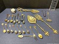 Full view of 24 assorted tableware items including collectible spoons, serving utensils, salt cellar, and sugar tongs.