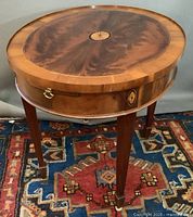 Full view of one oval side table showing burled veneer top, brass ring pull slide and tapered legs on area rug.