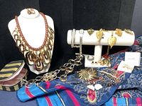 Overall lot showing multiple necklaces, brooches, earrings, bracelet and scarf spread on fabric background.