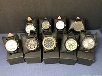 Front view of all 9 men's wristwatches arranged in two rows on black stands showing various dial designs and black leather bands.