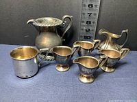 Seven vessels including a large creamer, four small creamers, a baby cup, and mini pitcher arranged with ruler for scale.