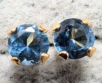 Close-up front angle showing one earring with round faceted blue gemstone in yellow gold prong setting against a soft fabric background.