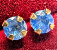 Close-up image of a pair of round blue sapphire stud earrings set in 10K yellow gold with four prongs each, displayed on red fabric.