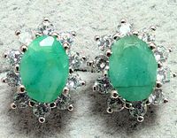 Two silver earrings with oval green emerald centers surrounded by clear cubic zirconia stones in floral halo design, laid side by side on gray background.