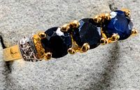 Close-up angled view of silver ring featuring three round blue sapphires in gold prong settings, with small clear accent stones on band near sapphires.