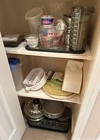 Kitchen shelf with assorted bakeware including glass and plastic measuring jugs, Pyrex glass mixing bowl, metal cake pans, muffin tray, and various other kitchen tools.
