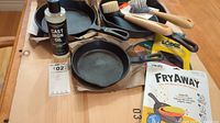 Cast iron fry pans and cleaning accessories including bottle of cast iron oil, brushes, scrapers, and FryAway packets on wooden surface.
