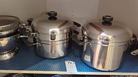 Large and medium stainless steel pots with lids, stacked steamer pot beside them on a shelf.