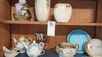 Image of assorted fine china and porcelain items on a shelf, including teapots, vases, and decorative plates.