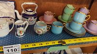 Demitasse cups and saucers in pastel colors with gold trim, along with some milk jugs and a teapot with floral and gold decorations arranged on a shelf. Measurement tape for scale.