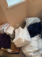Stack of various linens including white towels and assorted bed linens with floral patterns and dark colored fabric, placed near a window.