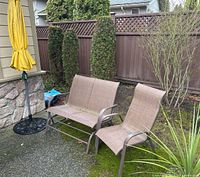 Photo of patio furniture including single rocker chair, two-seat patio bench rocker chair, and yellow patio umbrella closed and standing in weighted base outdoors.