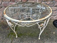 Photo of round patio table with clear glass top and rusted white metal frame on stone background, showing full table from an angle.