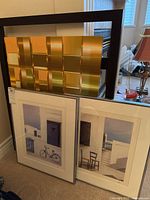 Two framed art prints leaning against a wall showing Mediterranean architecture scenes and a large abstract gold metallic tile design print stacked behind.